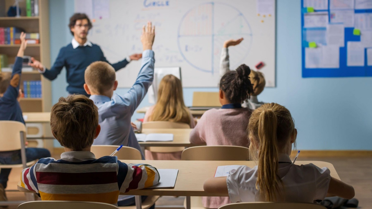 A classroom: A male teacher wearing a blue sweater stands in front of young children at their desks, some raising their hands A classroom: A male teacher wearing a blue sweater stands in front of young children at their desks, some raising their hands
