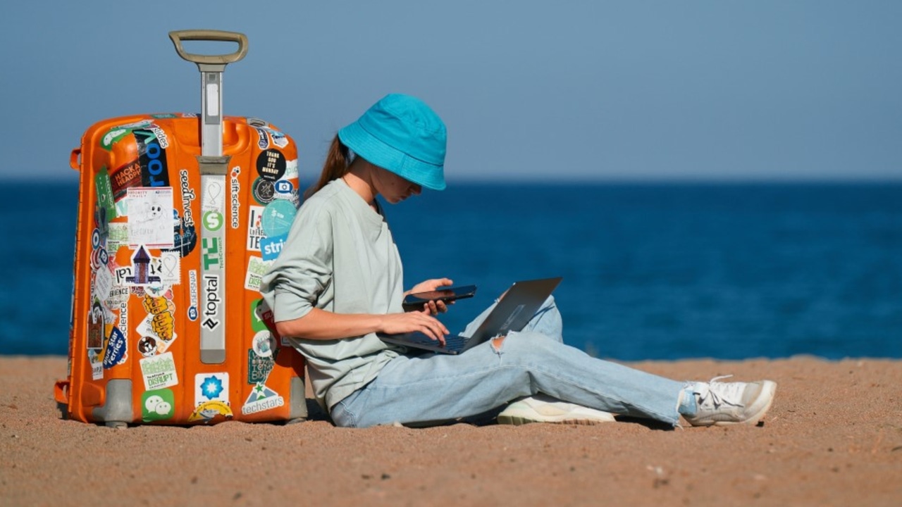 A person in a blue hat sitting on the beach with a suitcase and a laptop.
