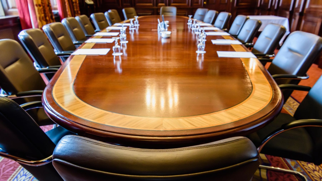 Wooden boardroom table; no one is seated but there are water glasses and paper on table Wooden boardroom table; no one is seated but there are water glasses and paper on table